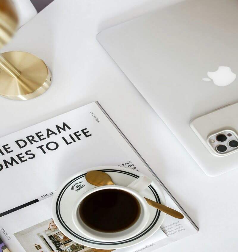 Minimalist workspace with a cup of coffee, a gold spoon, an Apple laptop, and a magazine titled ‘The Dream Comes to Life,’ symbolizing planning and achieving goals.
