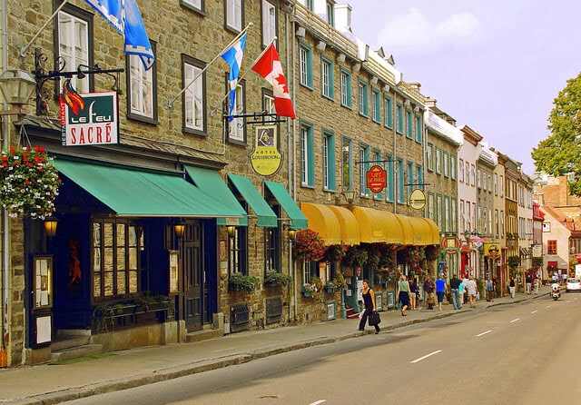 A lively street scene in Old Quebec City featuring historic stone buildings, awnings over storefronts, a Quebec flag, and a Canadian flag flying above people walking on the sidewalk.