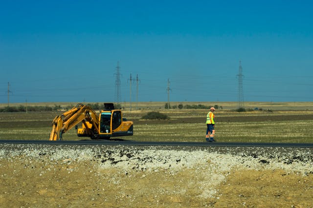 A construction worker in a high-visibility vest walks along a rural roadside near an excavator in an open field under a clear blue sky.